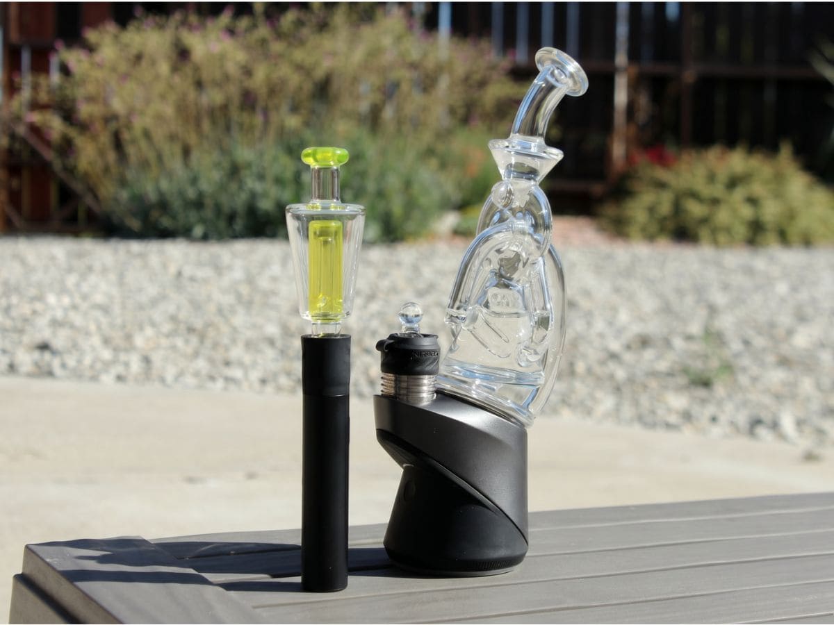 A Puffco Peak with a clear recycler glass top beside a second Puffco with a yellow glass attachment, outdoors on a deck